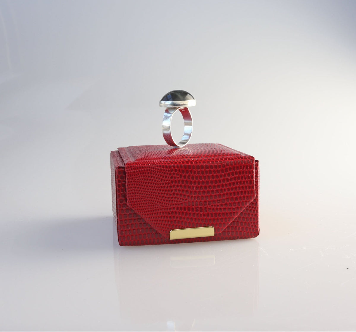A Blue Tiger’s Eye gemstone ring handcrafted in solid Argentium 935 silver with fine silver bezel, displayed on top of a small red textured envelope-style jewelry box with a gold clasp, set against a white studio background.