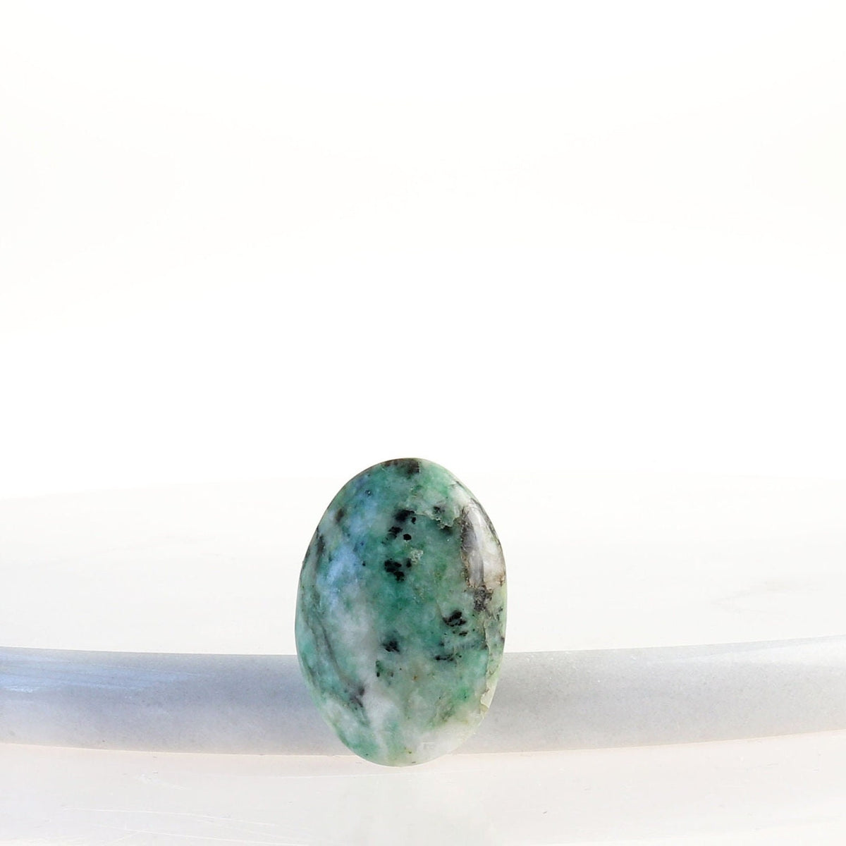 Front view of African Turquoise Jasper cabochon standing upright showing its polished surface and natural  blue-green, teal, cream and black patterning.  Shown against white marble slab against white background.