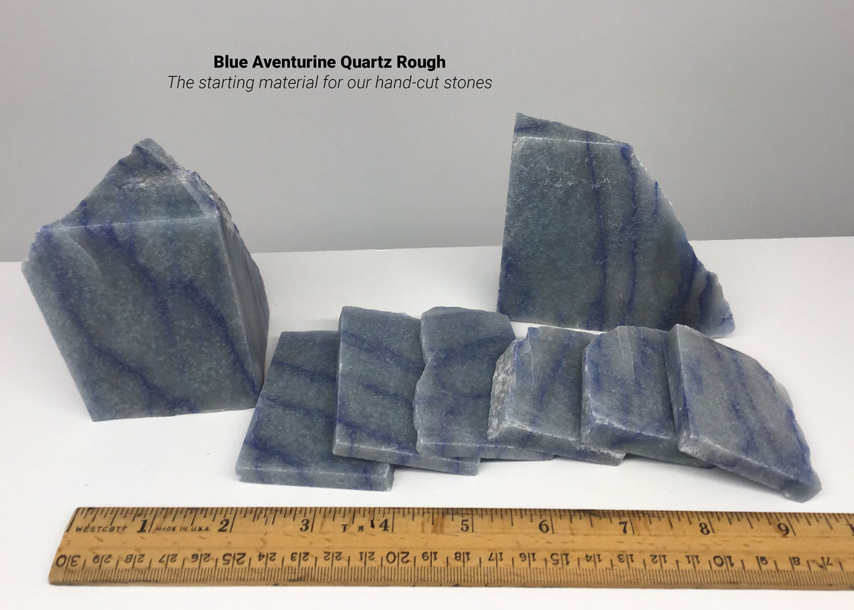Blue Aventurine Quartz rough and slabs used for our hand-cut gemstone jewelry. Arranged on a white surface with a wooden ruler at the bottom for scale, with text reading 'Blue Aventurine Quartz Rough The starting material for our hand-cut stones'.