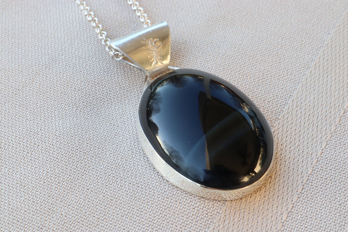 An oval natural Blue Tiger’s Eye pendant in a polished Argentium 935 silver bezel, suspended from a silver chain and resting on a textured beige fabric background.