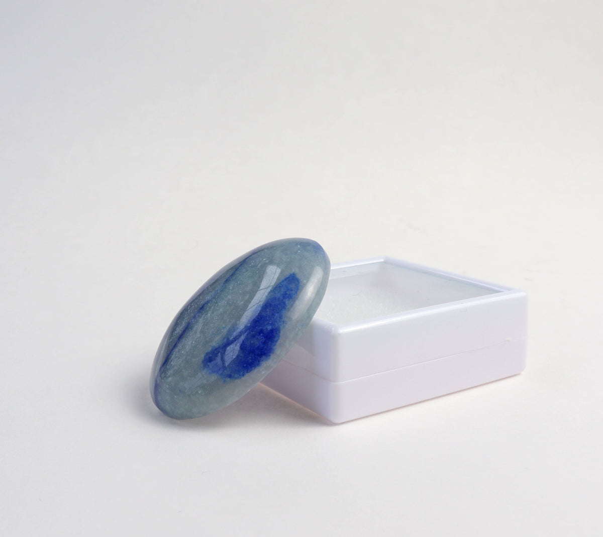 A polished blue Aventurine and white stone cabochon resting on its white plastic display box, set against a seamless light gray background.
