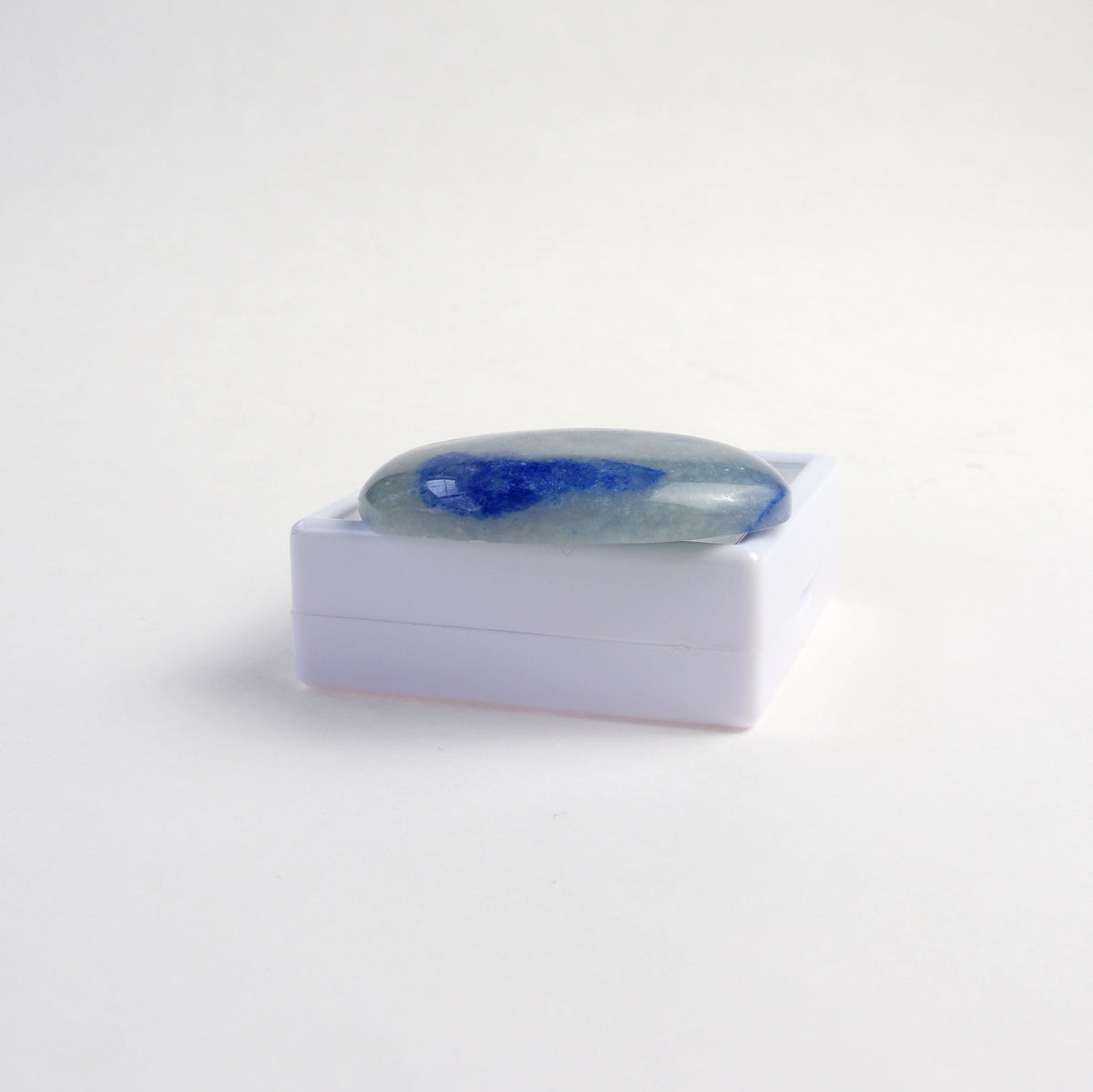 A polished Blue Aventurine oval shaped cabochon resting on a small white gift gem box, set against a  white background.