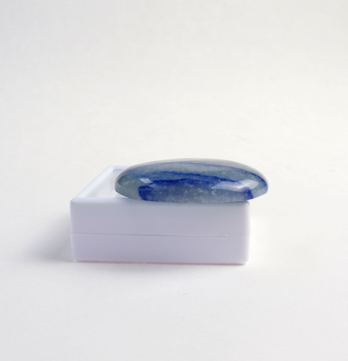A polished Blue Aventurine oval shaped cabochon resting on a small white gift gem box shown opposite side view of stone, set against a  white background.