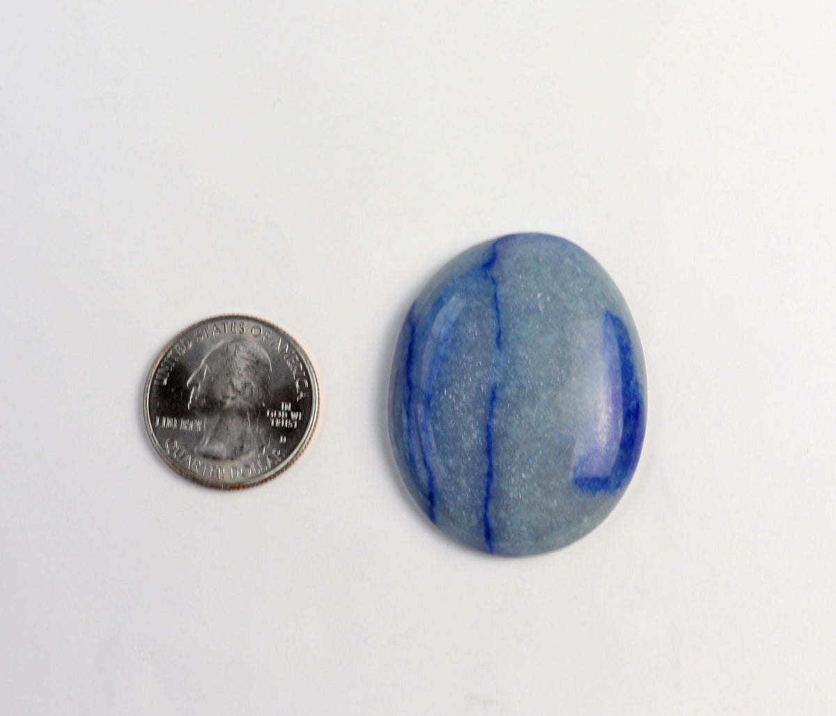 A polished blue oval Aventurine gemstone with natural banding, shown next to a US quarter coin for scale on a plain white background.