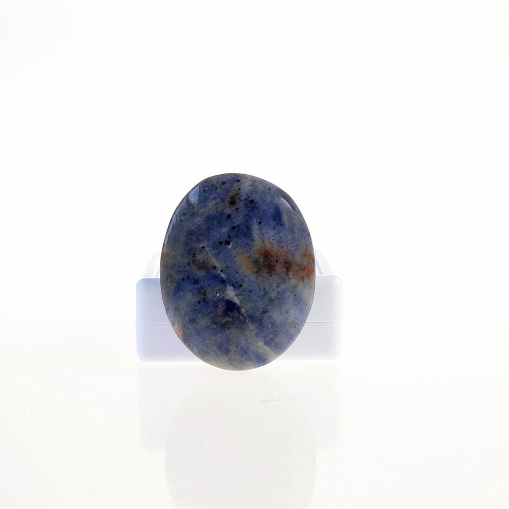 Hand-cut Blue Foidolite Sodalite oval cabochon with mottled blue, creamy white, gray, and warm brown natural patterns.  Shown against a white gem box against a white background.