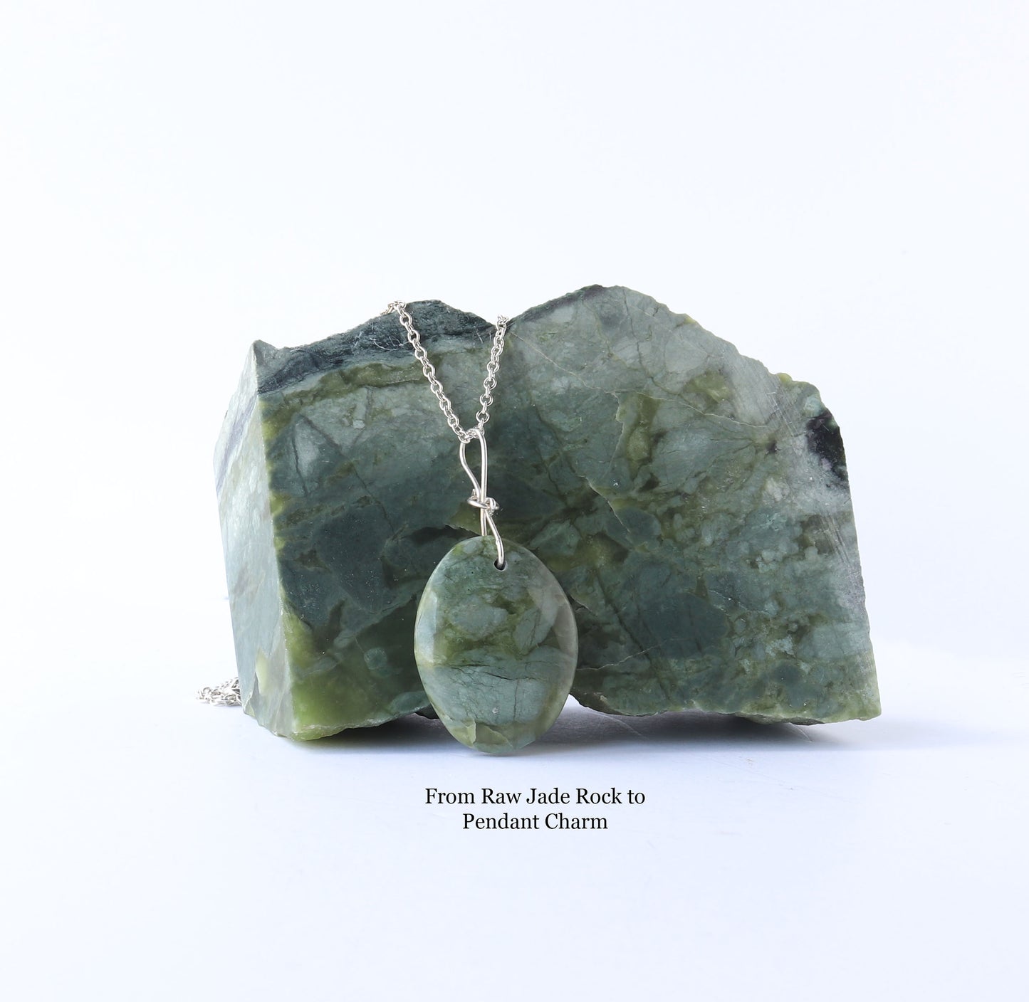 A polished California jade oval pendant charm suspended from a silver chain and displayed on top of the raw jade rock  against a white background. Text below reads 'From Raw Jade Rock to Pendant Charm'.