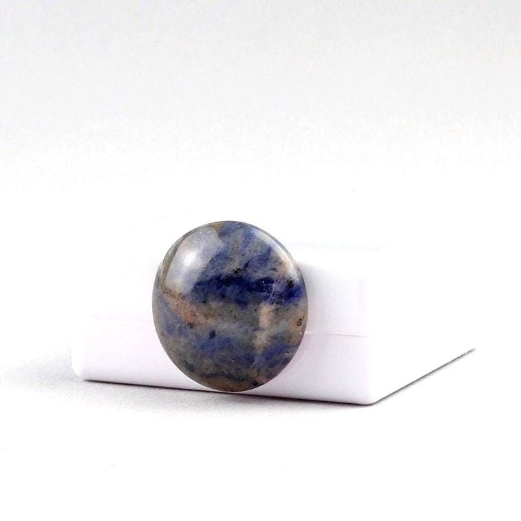 A single round Blue Foidolite Sodalite cabochon hand-cut 19.64 mm with a marbled pattern of deep blue, slate gray, and brown, standing next to a white gem box and photographed on a clean white background.