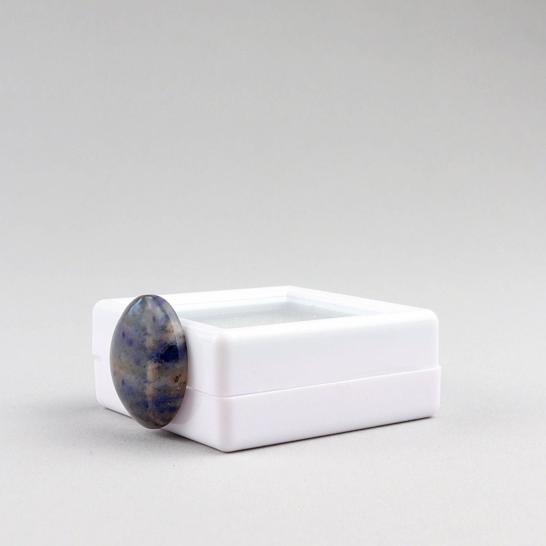 A side angle of a single round Blue Foidolite Sodalite cabochon hand-cut 19.64 mm with a marbled pattern of deep blue, slate gray, and brown, standing next to a white gem box and photographed on a clean white background.