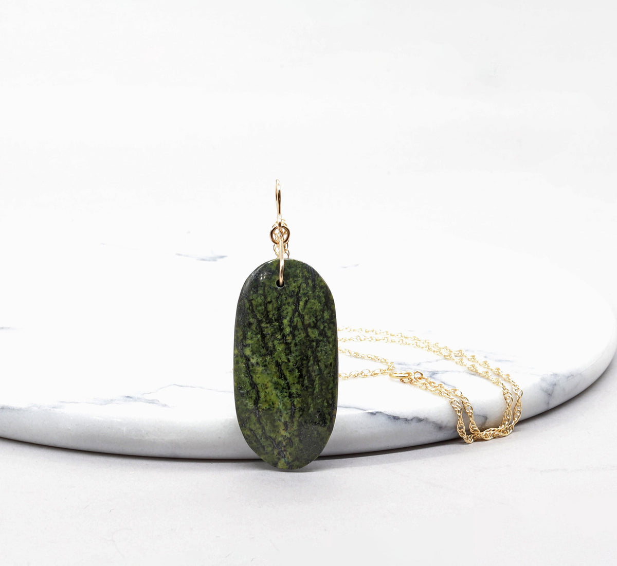 A Forest Green Jasper pendant necklace with a bail in gold-fill and a rope style gold-filled chain, displayed on a round white marble slab against a white background.