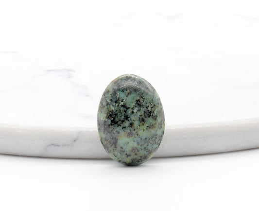 A Fraser River polished gemstone with a mottled green and black matrix pattern, resting on a white marble slab against a white background.