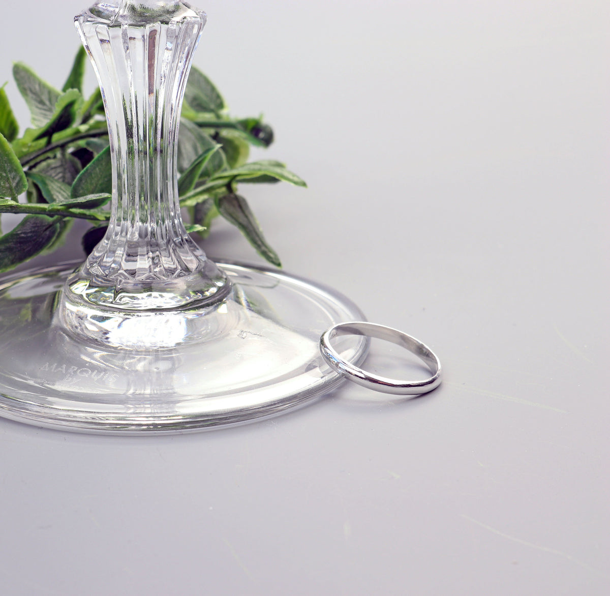 Handcrafted solid Argentium 935 silver half-round ring with smooth polished band displayed beside a clear glass pedestal. displayed against a gray background with a glass base and green leaves blurred in the background.