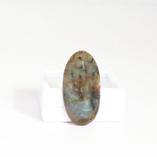 Natural Labradorite elongated oval cabochon with blue teal flash standing against white gem box shown on white background. 