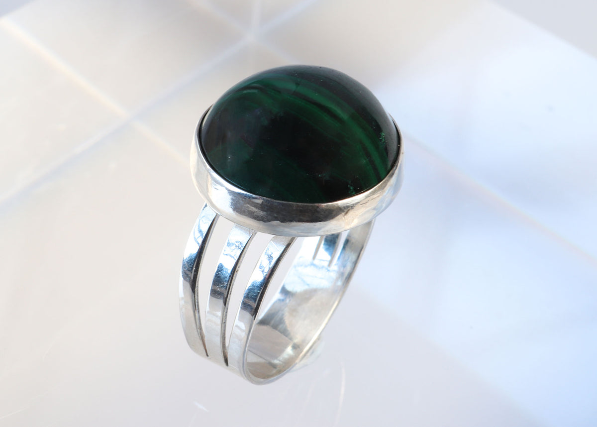 Handcrafted natural green Malachite ring with oval cabochon, polished fine silver bezel and solid Argentium 935 silver split-shank band. Shown standing on an acrylic block against light gray background.