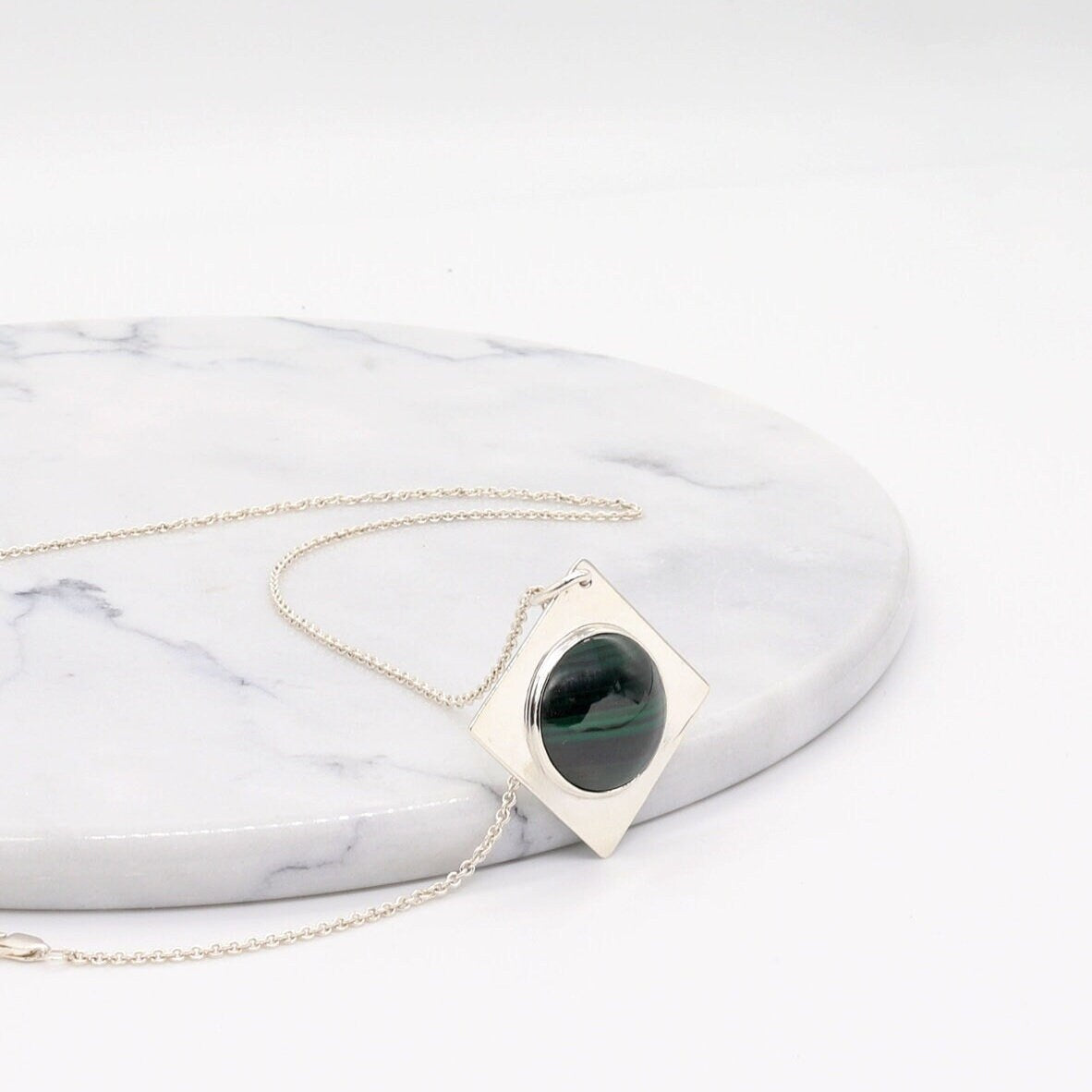 Handcrafted malachite pendant necklace in Argentium silver with geometric setting shown against white marble slab against a white background.