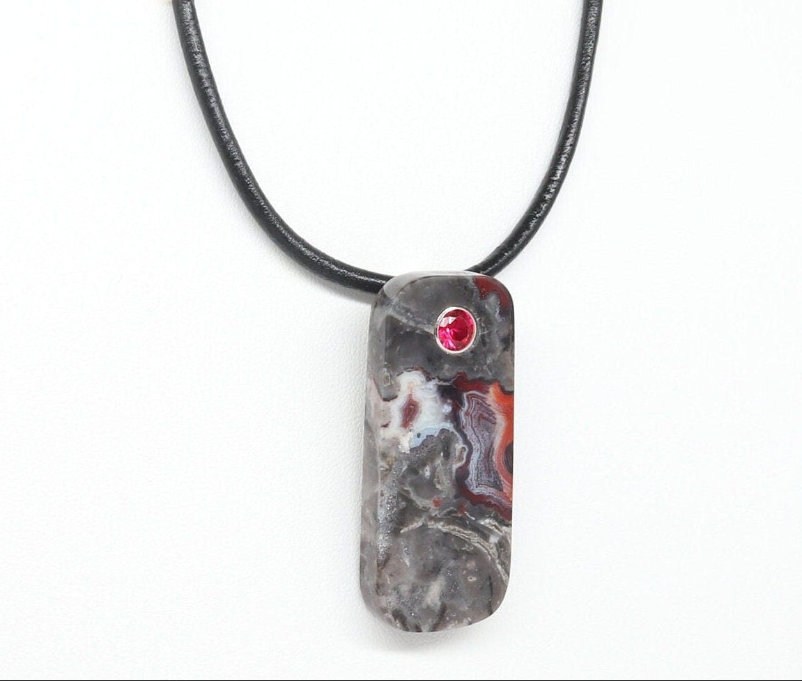 A 2 inch tag shaped Mexico Agate, handcut and freeform polished with a 6mm lab ruby inlay. The pendant has been fashioned with a bail in premium 935 Argentium® Silver, resistant to tarnishing and its brilliant luster. 