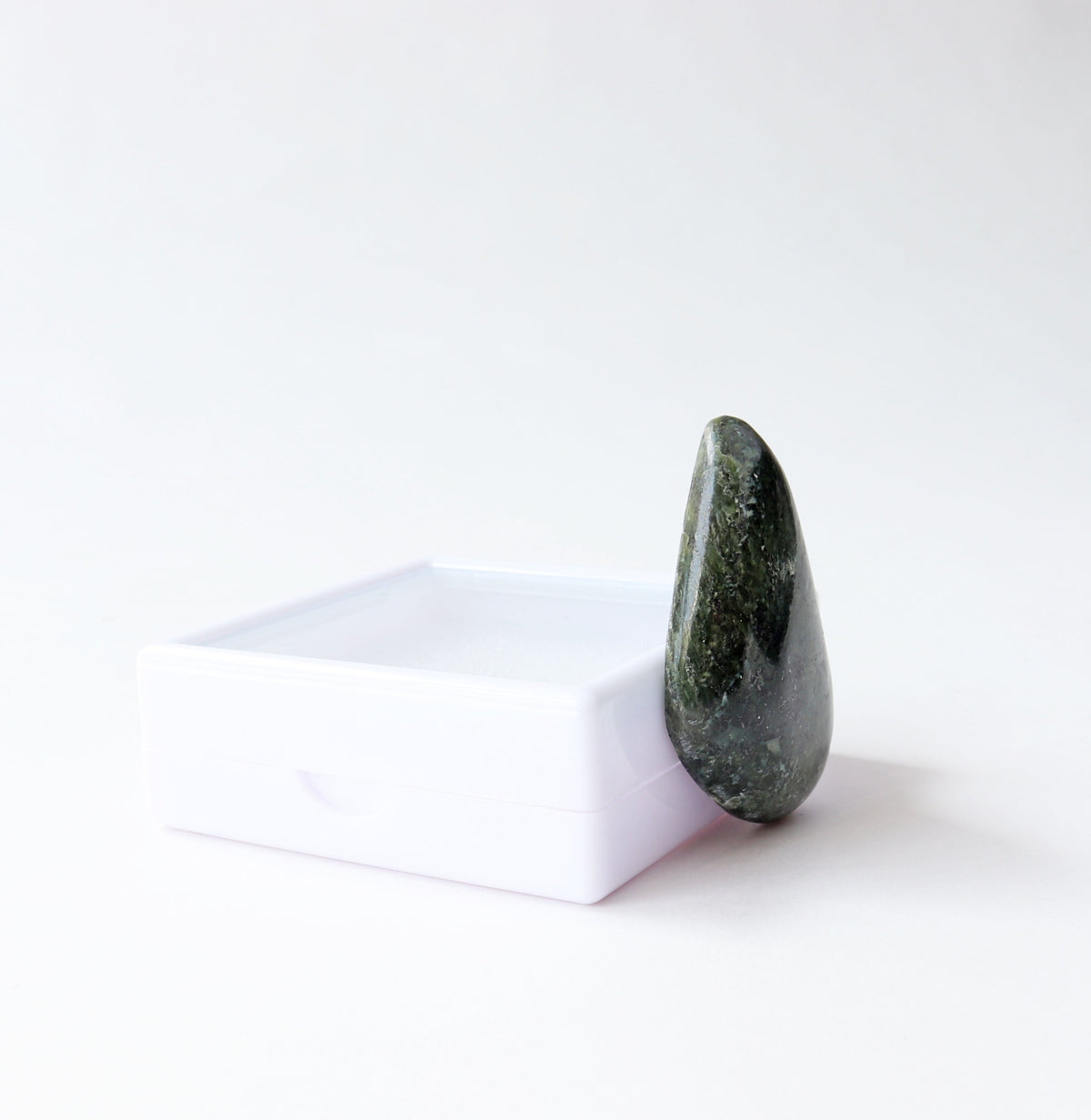 A freeform polished dark green Moss Agate cabochon standing upright at a left angle against a small white rectangular block on a clean white background.