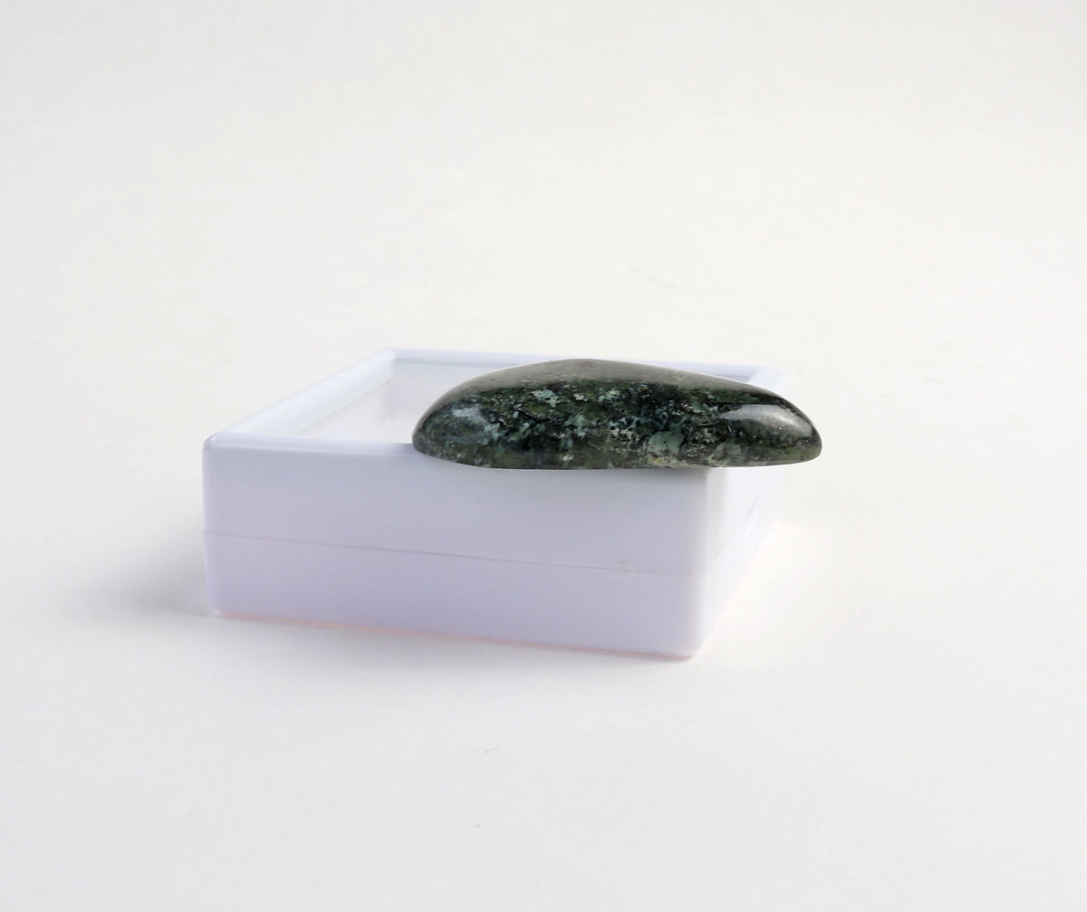 A side-view of this freeform polished dark green Moss Agate cabochon, displayed on top of a small translucent white square box against a clean white background.