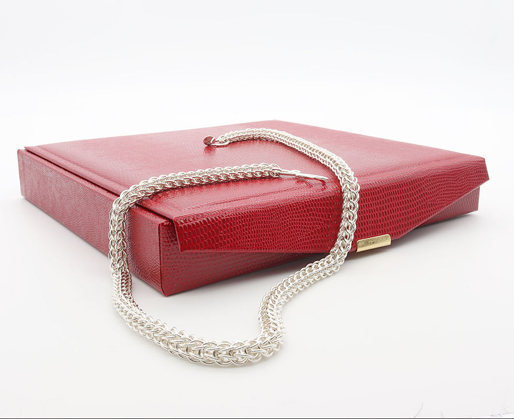 A red textured leather gift box with a gold color clasp with a handmade Argentium silver chain persian weave necklace draped over the corner of the box , resting on a white background.