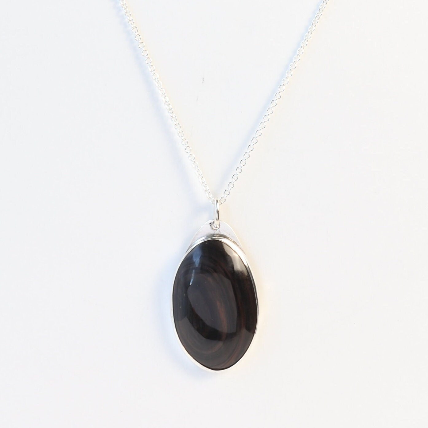 A Mahogany Obsidian pendant displays swirling deep reddish-brown and dark mahogany hues, set in  Argentium® 935 Silver shown on white background with silver necklace.