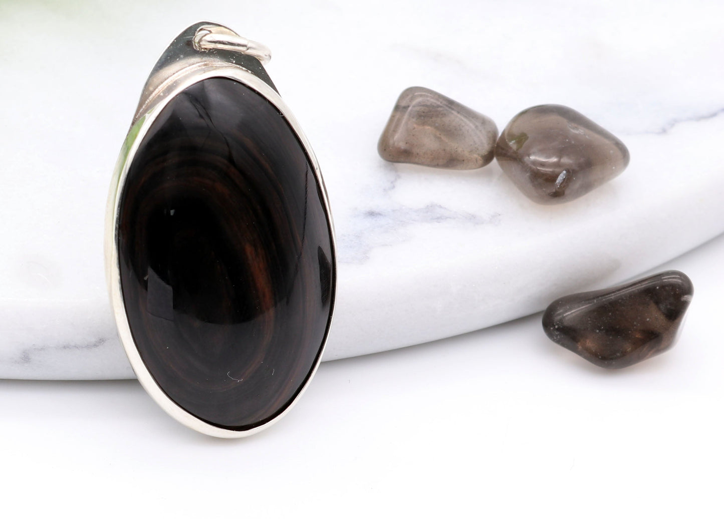 A Mahogany Obsidian pendant with swirling patterns in deep reddish-brown and dark mahogany hues,  set in Argentium® 935 Silver.  Shown on white background next to raw mahogany obsidian pebbles for props.