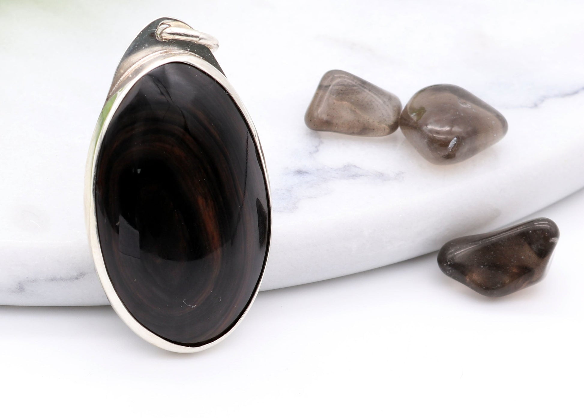 A Mahogany Obsidian pendant with swirling patterns in deep reddish-brown and dark mahogany hues,  set in Argentium® 935 Silver.  Shown on white background next to raw mahogany obsidian pebbles for props.