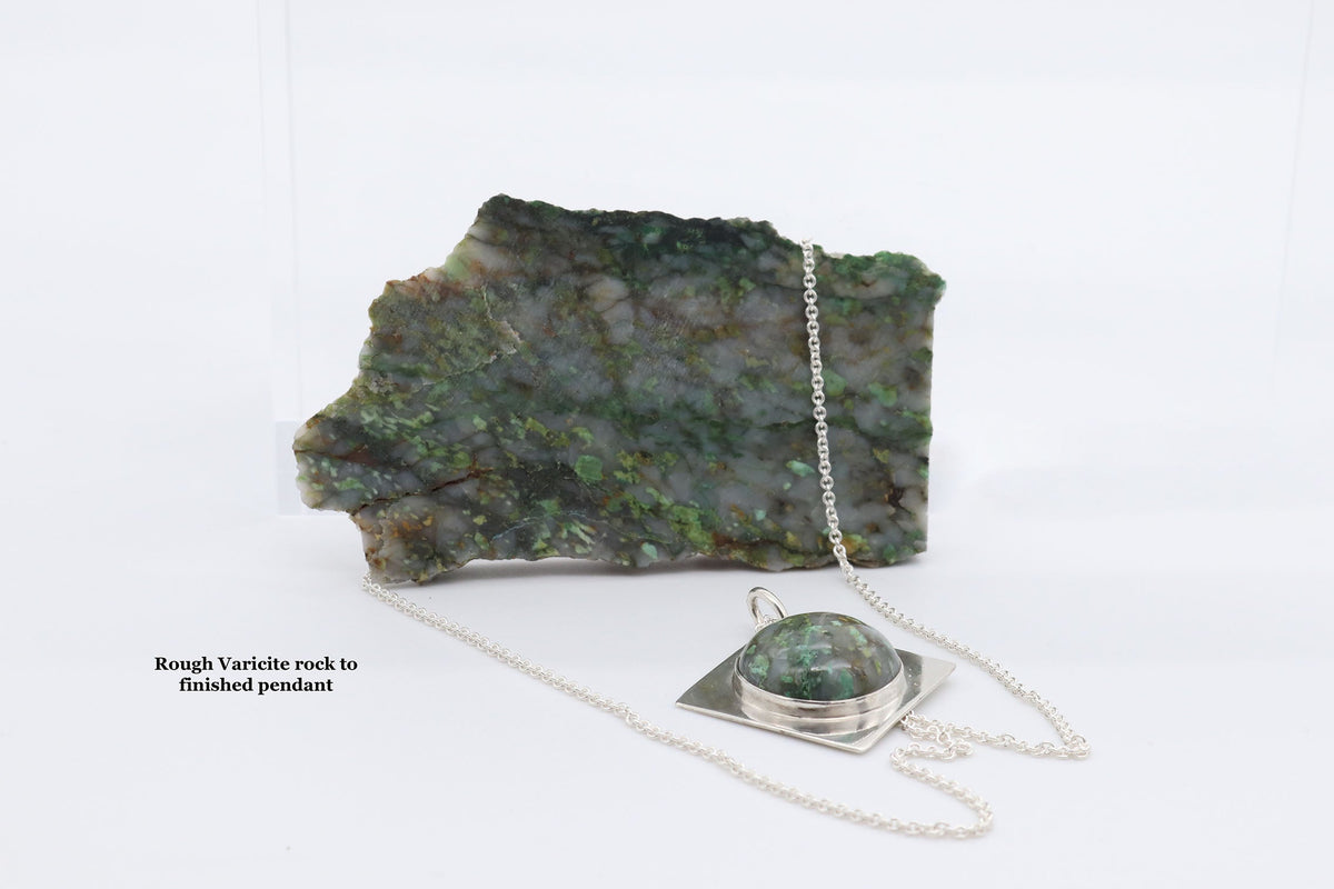 A silver necklace with a polished green varicite stone pendant displayed on a rough green varicite rock specimen, with a small text label reading 'Rough Varicite rock to finished pendant' on a white background.