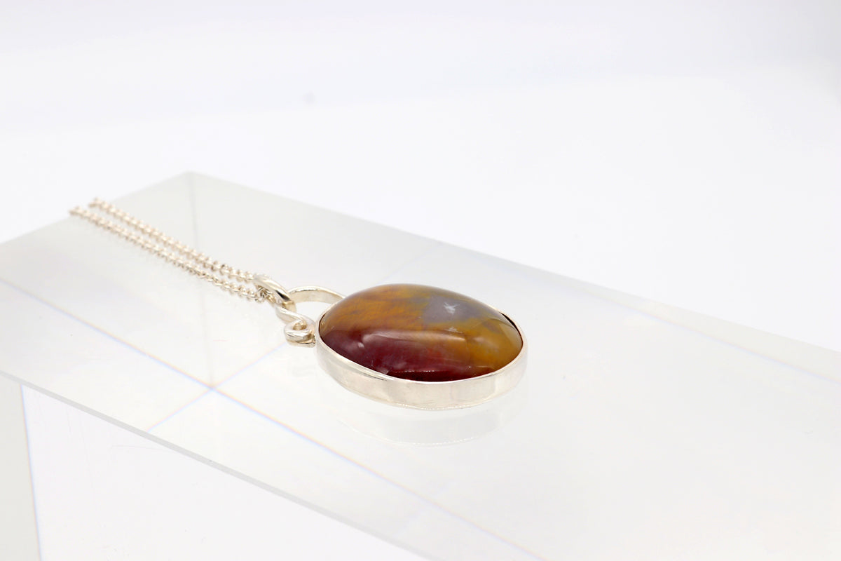 A silver chain necklace with an oval Petrified Rainbow Wood pendant featuring warm golden, rust, gray, and earthy tones along with the distinctive patterns preserved in the stone, resting on a clear acrylic display block against a white background.