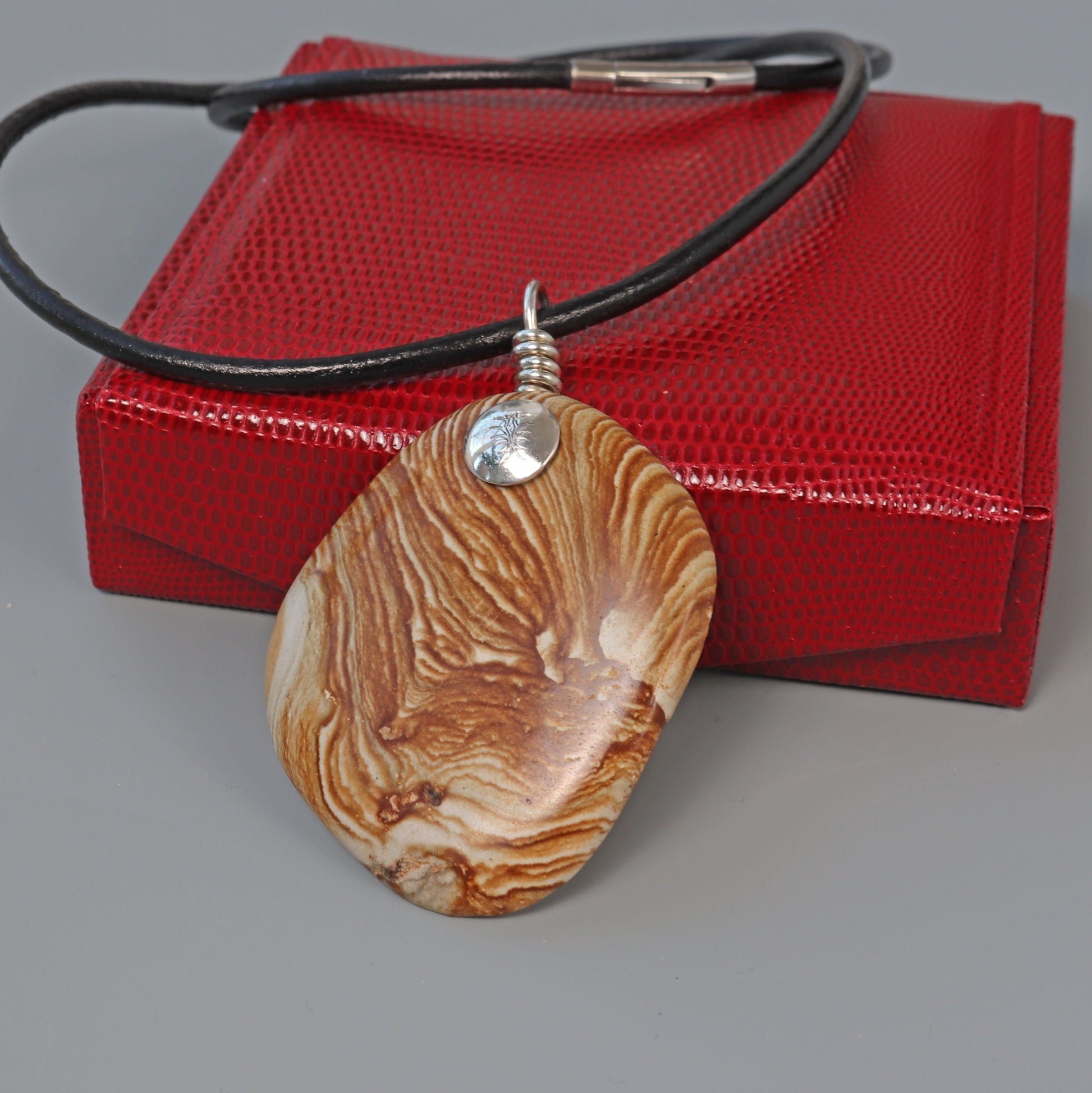 A natural 2.5inch Picture Jasper gemstone, handcut and polished on both sides to showcase the earthy tones, warm browns, creamy beiges, and nature's intricate patterns.  The custom handcrafted coil bail is from sterling Argentium® 935 Silver renowned for its resistance to tarnishing and its durable quality.
To complete the look a complementary 3 mm Leather Cord Necklace is included.  Ready to gift in a red faux leather gift box.