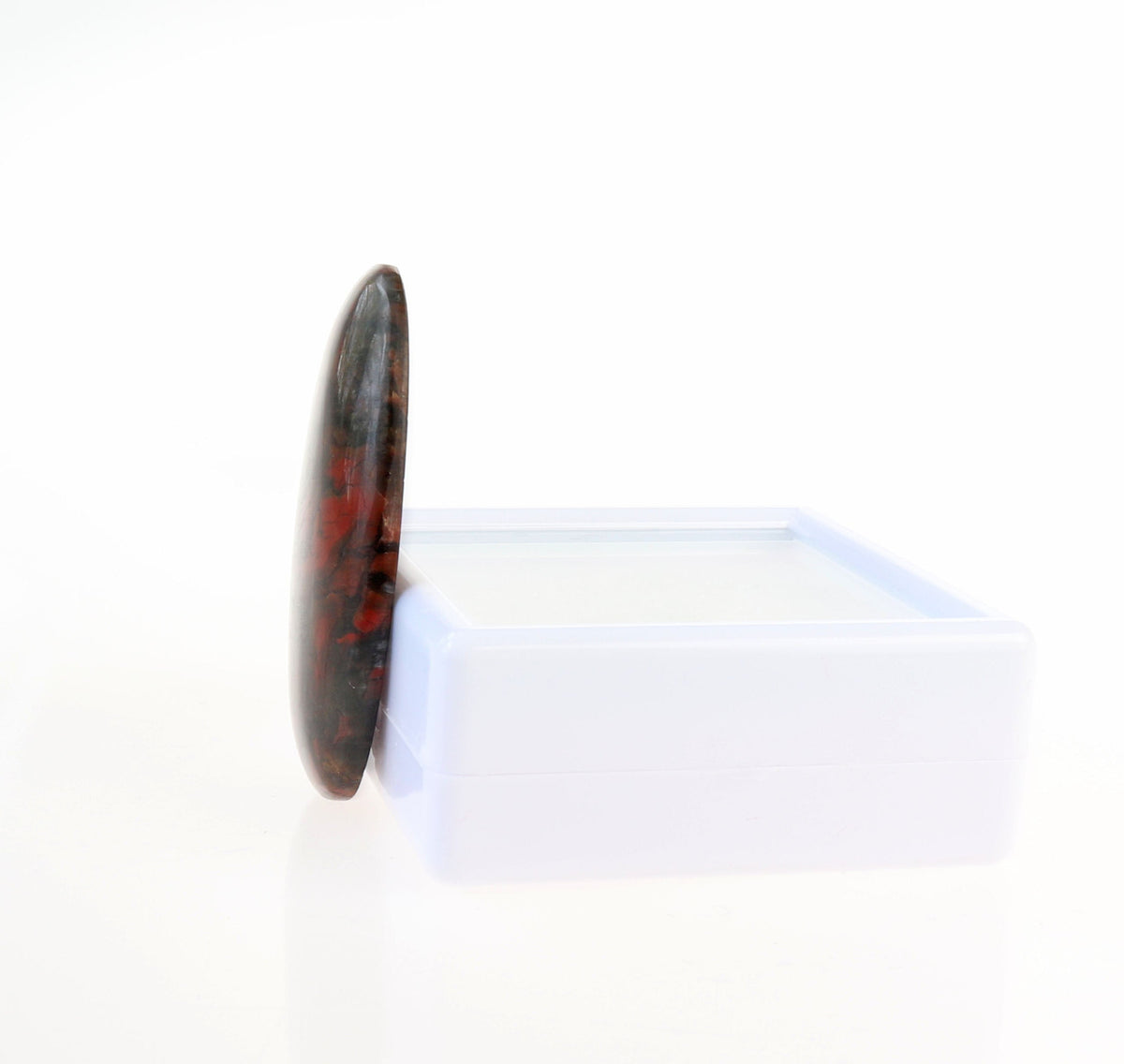 Side view of Natural Red and Black jasper gemstone cabochon, hand-cut and polished into an oval shape to reveal natural mottled patterns of rich reds, earthy blacks and a small white inclusion within the stone.  Shown against white gem box on white background.