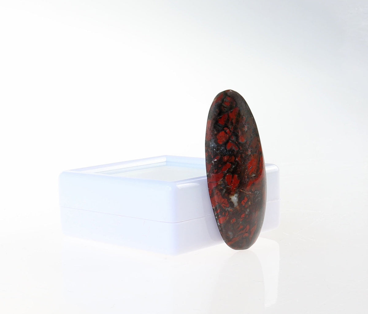 Side left angle view of a Natural Red and Black jasper cabochon, hand-cut and polished into an oval shape to reveal natural mottled patterns of rich reds, earthy blacks and a small white crystal inclusion within the stone.  Shown against white gem box on white background.