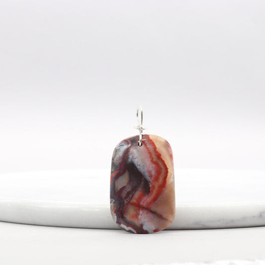 A 1.95 inch Red Mexico Agate hand-cut and freeform polished to bring out the mauve gray and deep red patterns and translucent colors. Crafted with an original infinity bow wire bail in Argentium 935 silver known for its higher silver content and its ability to resist tarnishing and keep on shining.