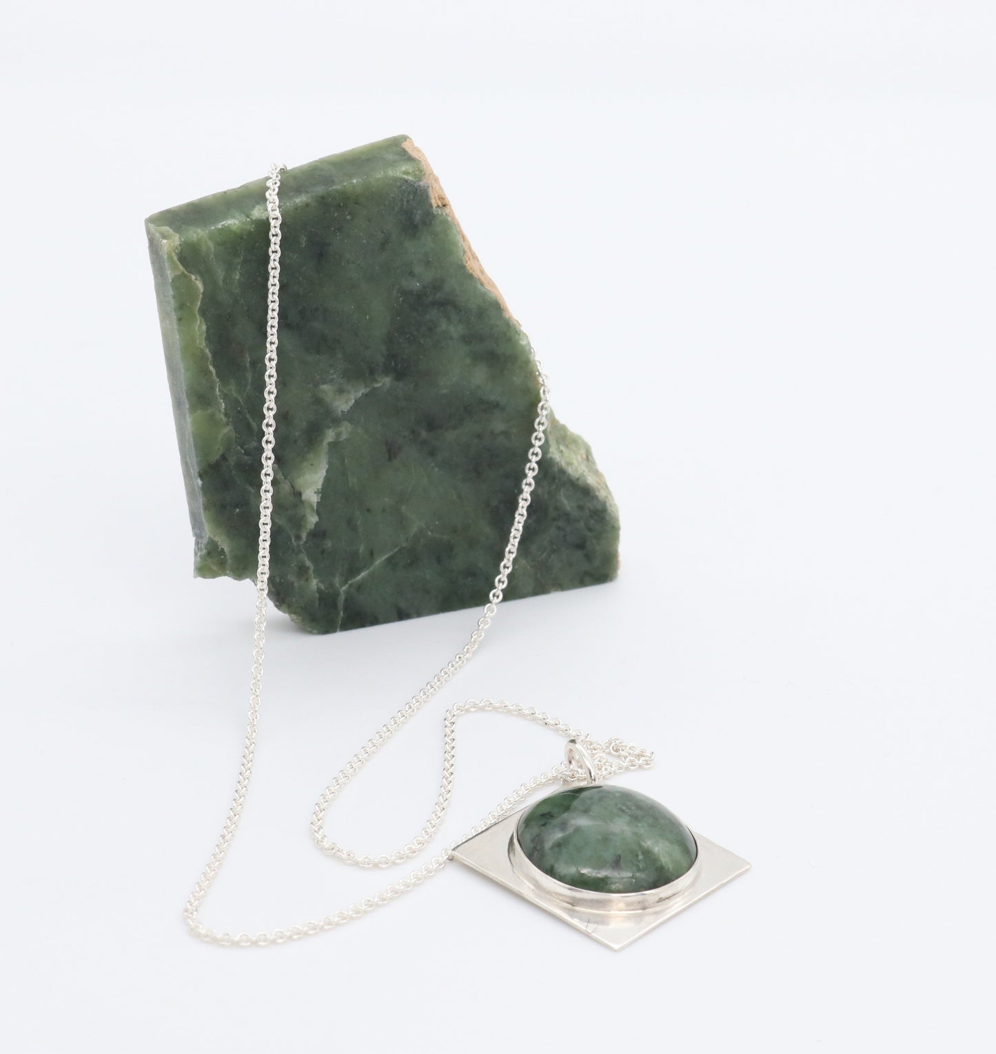 A silver necklace with a square pendant featuring a Wyoming cabochon stone, set in solid Argentium silver displayed next to rough Wyoming jade rock used to craft this pendant against white background.