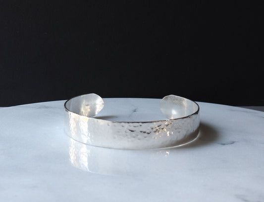 Hand-hammered sterling silver cuff bracelet with a softly curved profile, resting on a light marble-patterned surface against a solid black background.