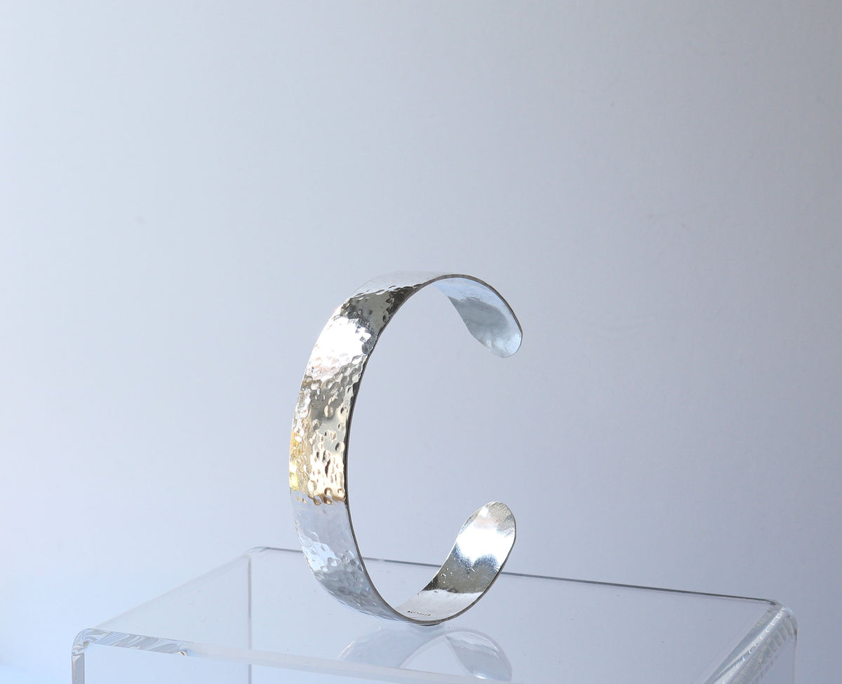 Hand-hammered sterling silver cuff bracelet with a softly curved profile displayed on a clear acrylic stand against a light gray background.
