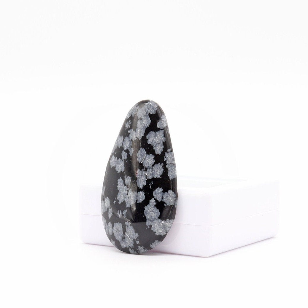 A one-of-a-kind Snowflake Obsidian cabochon, hand cut and polished.  Shown at an angle on a white background leaning on a white gem gift box.