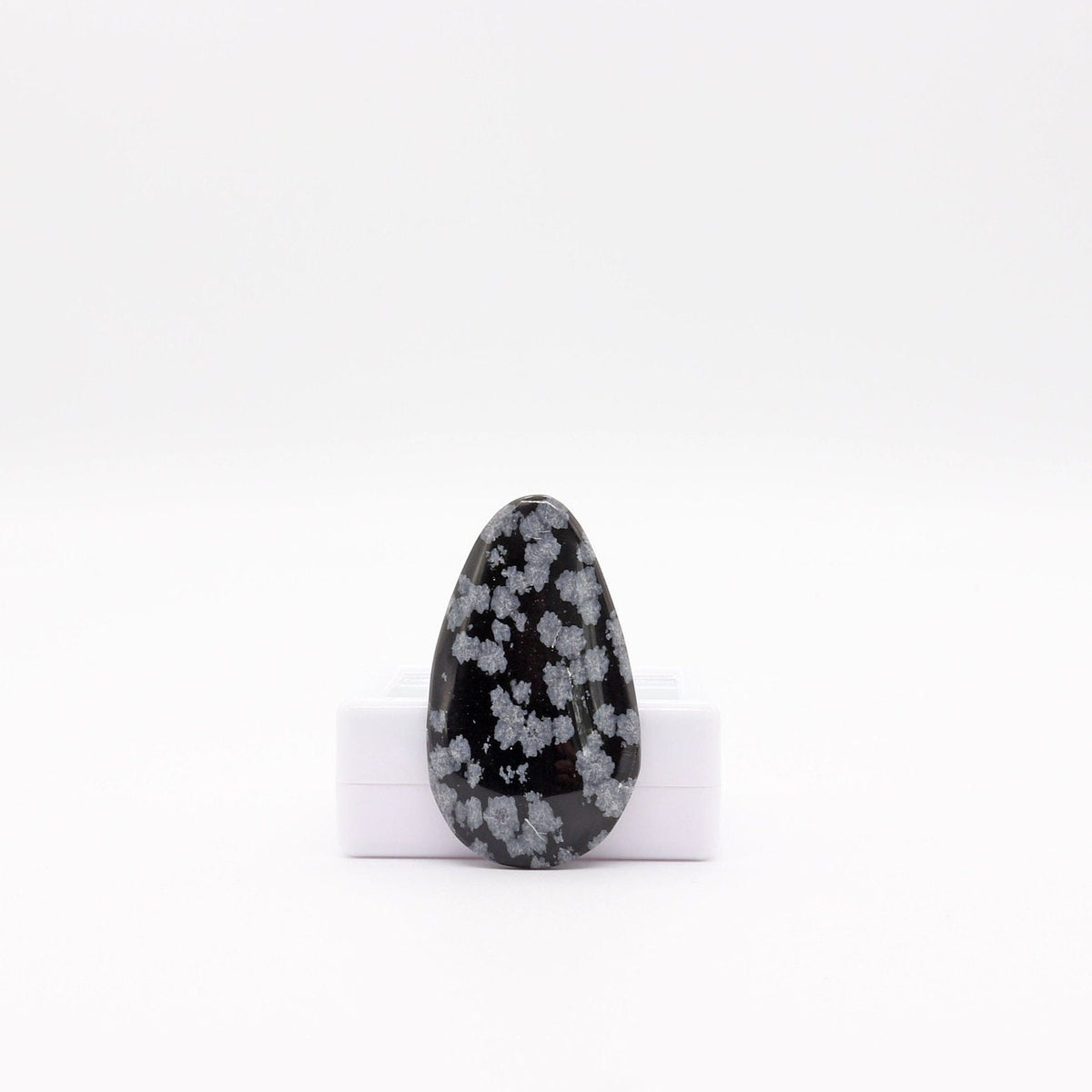 A one-of-a-kind Snowflake Obsidian cabochon, hand cut and polished in our Missouri lapidary studio.  Shown on a white background leaning on a white gem gift box.