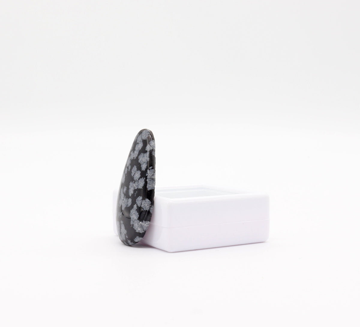 A teardrop-shaped Snowflake Obsidian with a mottled black and white pattern, standing upright in a side view on a small white gem gift box against a plain white background.