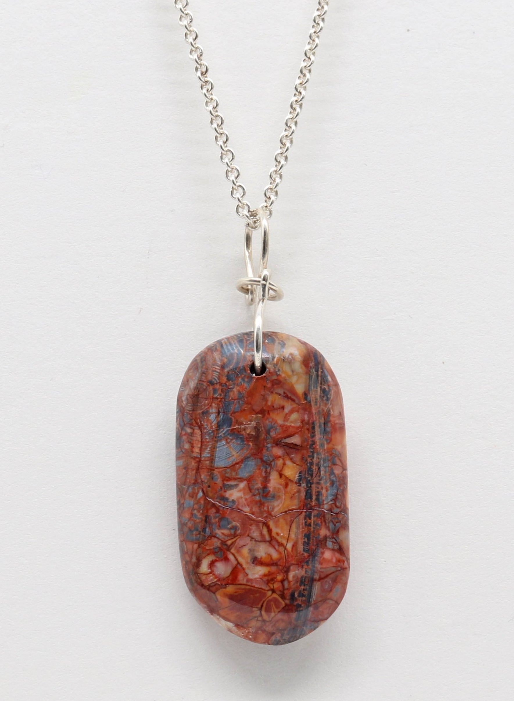 A Stony Creek Jasper gemstone pendant. Handcut and polished on both sides to showcase the unique red and blue patterns of the stone. &nbsp;The pendant is designed with an original infinity bow shaped wire bail made from Argentium® 935 Silver Shown on white background.