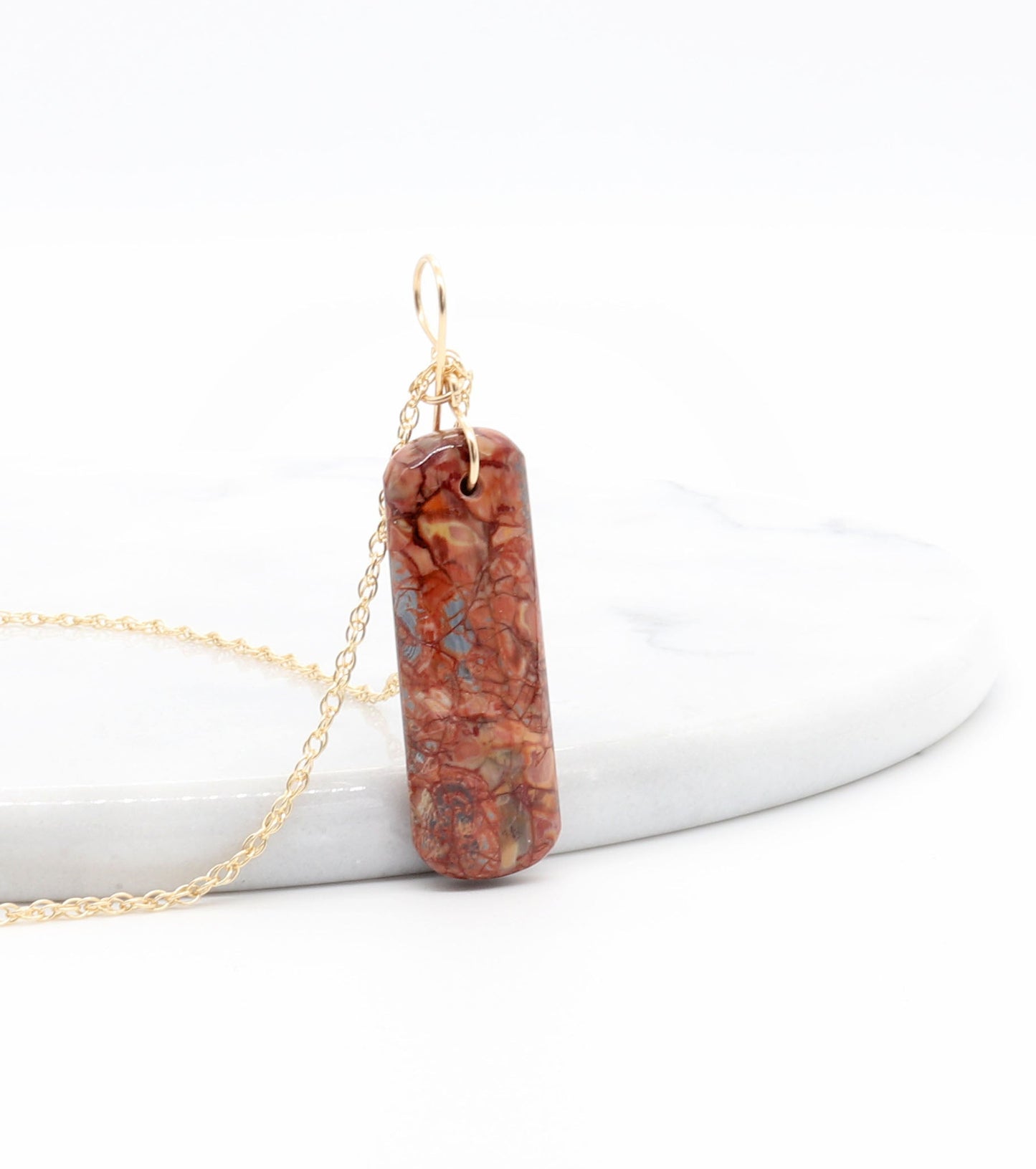 Side view of this Stony Creek Jasper pendant  hand-cut and polished features bold red and deep blue patterns suspended from a handcrafted 14/20 Yellow Gold-Filled Infinity Bow Bail with optional Gold-Filled Rope chain against white marble background.