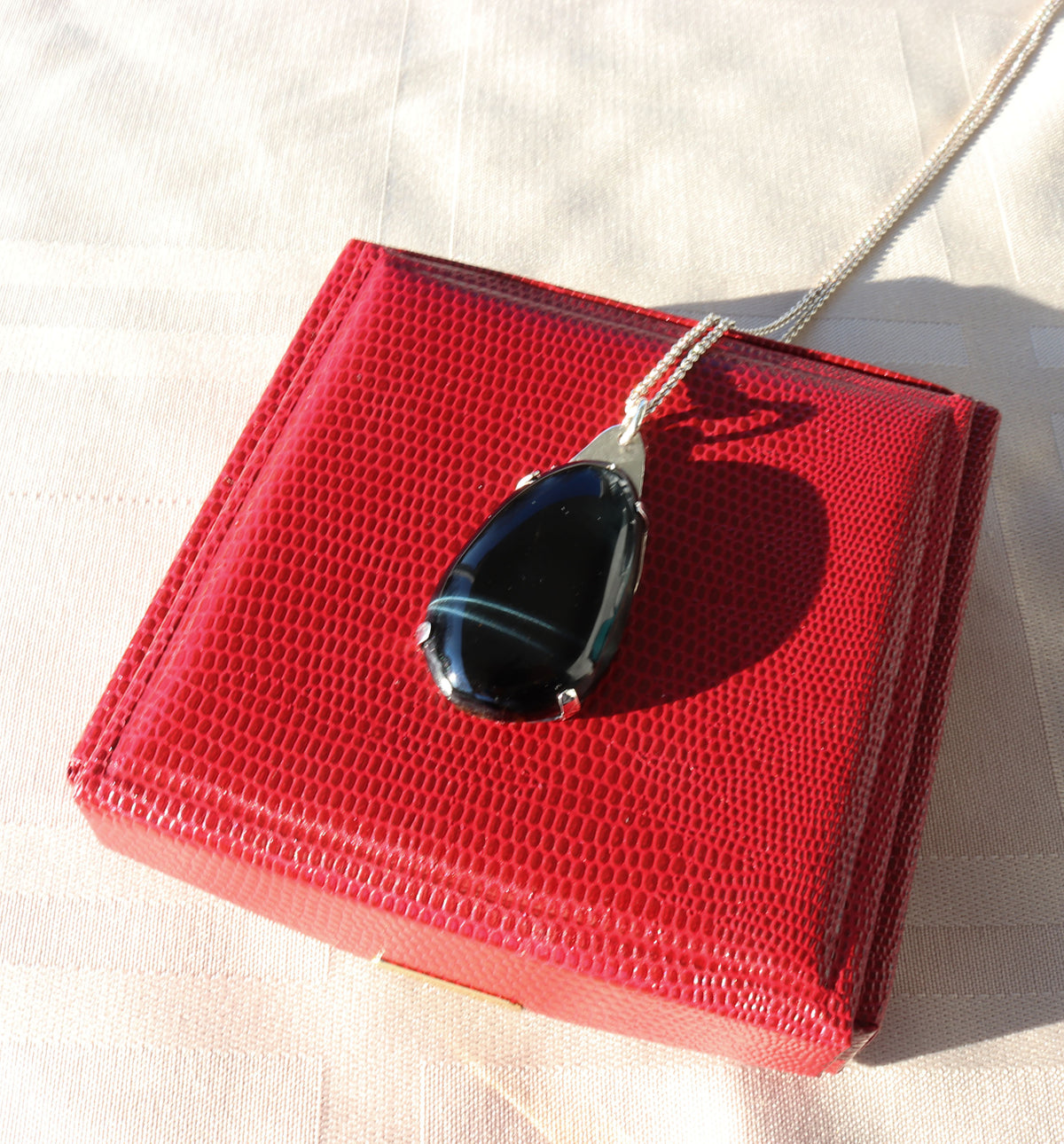 A Handcrafted African Blue Tiger's Eye teardrop pendant with deep blue-black reflective gemstone, solid Argentium 935 silver tabbed setting and silver chain, displayed on top of a textured red box against a light beige fabric background.