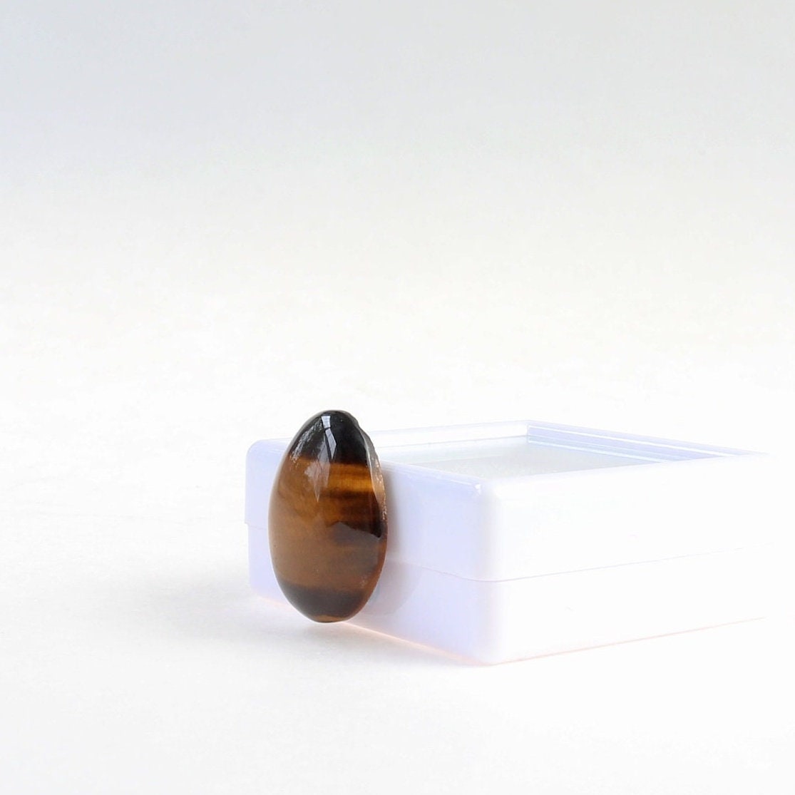 Hand-cut natural Tiger's Eye cabochon with golden-brown banding and chatoyancy, displayed at right angle against a white gem box.