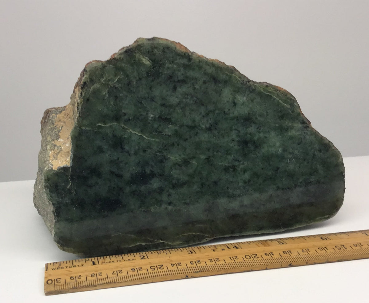 A rough Wyoming Jade stone with a natural rough texture, displayed on a white surface with a wooden ruler underneath for scale, against a plain light gray background.