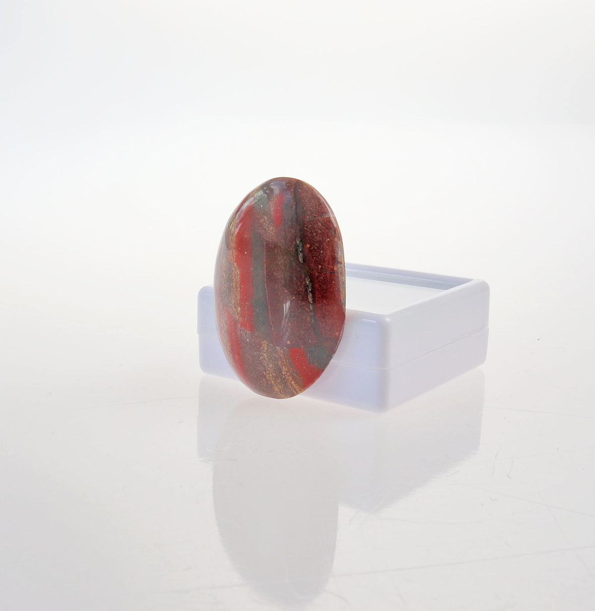 A polished oval Snakeskin Jasper stone with rich red and rust tones, accented by earthy brown, golden cream, and subtle gray areas, displayed on a small white stand against a clean white background.