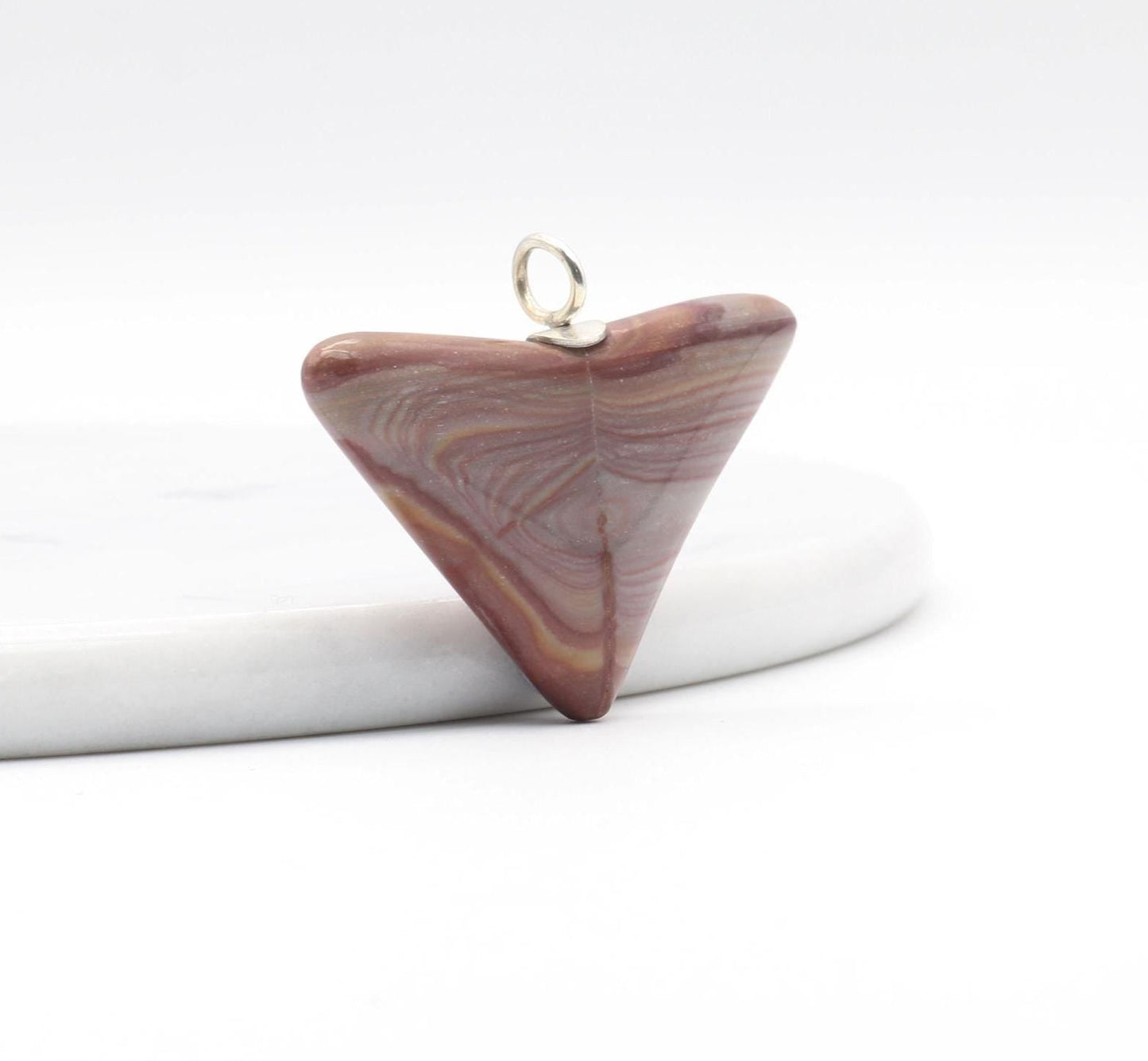 Angle view of this handcut polished Butterfly Shaped Petrified Rainbow Wood pendant featuring earthy tones of deep crimson, purple, and tangerine with handcrafted Argentium 935 silver bail shown against white marble slab and white background.