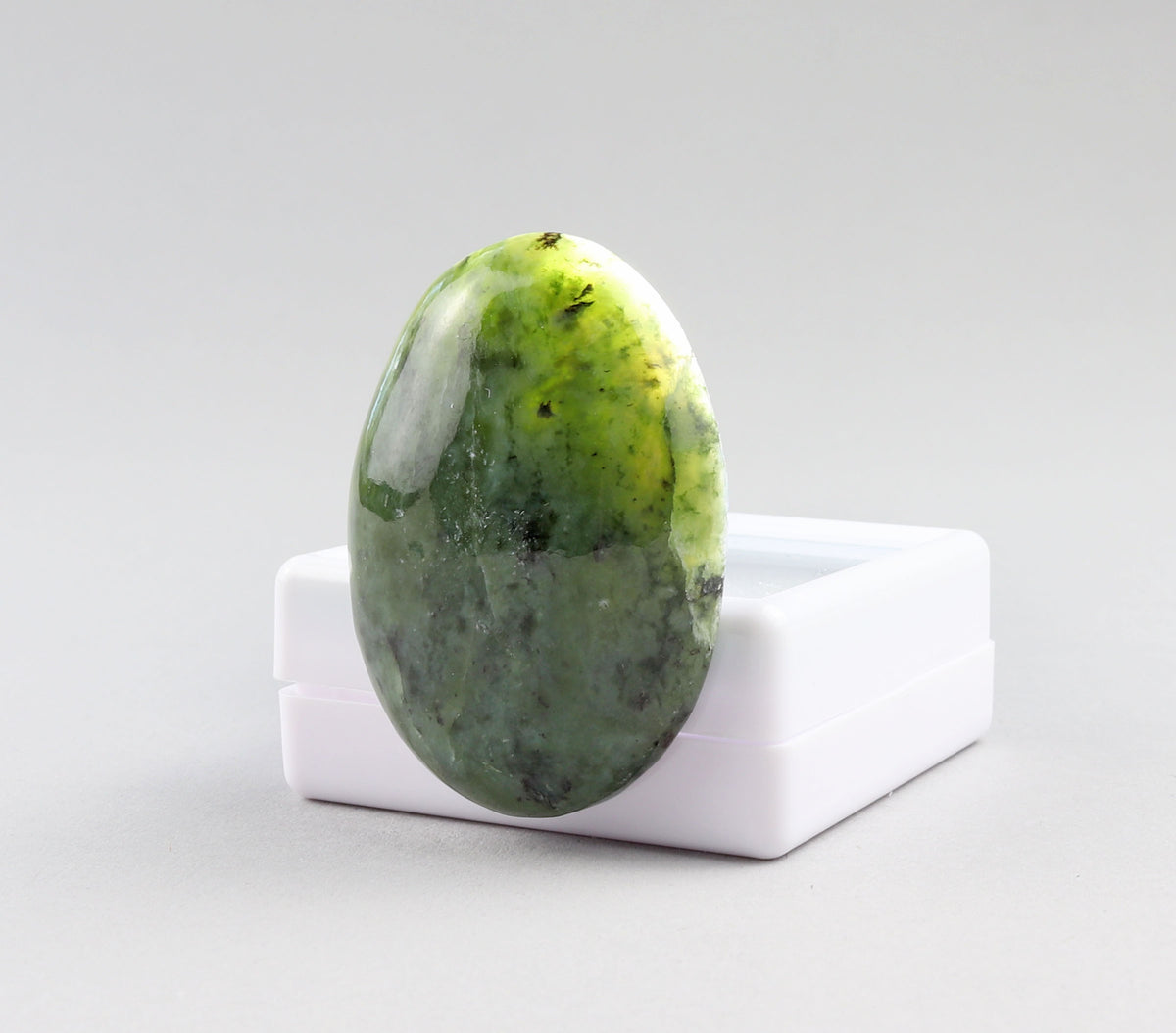 Backlit hand-cut oval Wyoming Nephrite Jade cabochon showing its translucent green tones and dark natural patterning, leaning against a white gem box on a white background.
