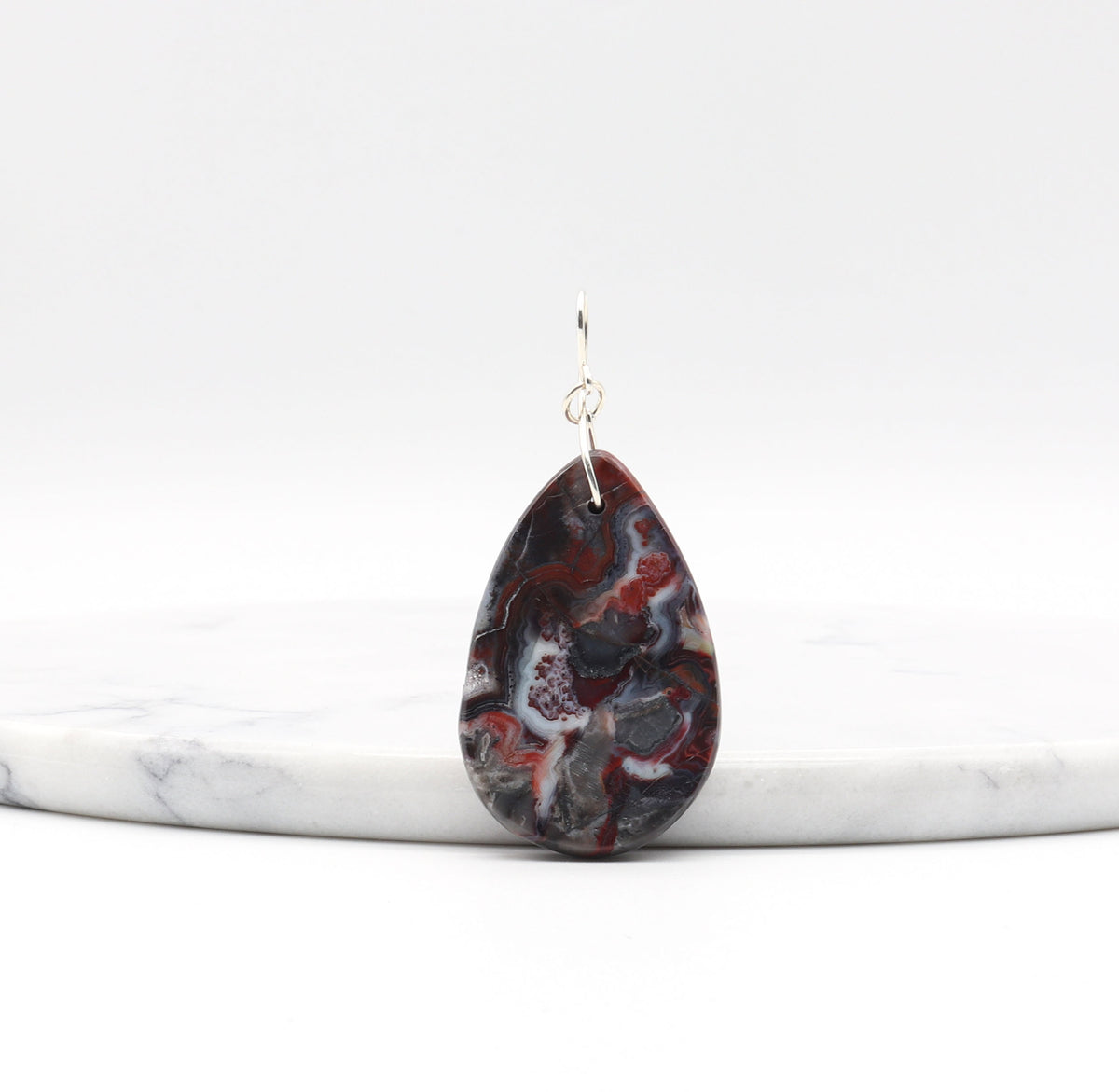 A back view of this teardrop-shaped Natural Mexican Red Agate druzy suspended from a silver wire hook and displayed on a white marble riser against a white background.