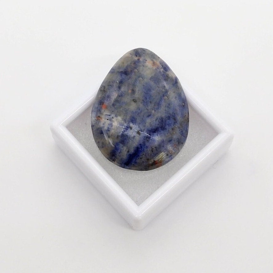 A one-of-a-kind Blue Foidolite teardrop cabochon, hand-cut and polished in our Missouri studio to showcase its natural blue, gray, cream, and subtle rust-colored patterning.  Shown leaning back on a white gem box against a white background.