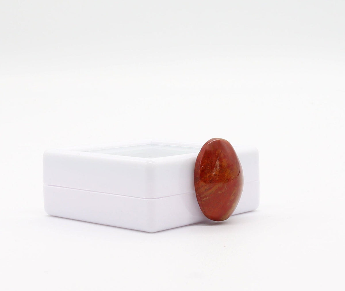 Left view of  Sonoran Sunrise Chrysocolla Cuprite gemstone, has been carefully handcut and polished into an oval shape cabochon, revealing its natural vibrant shades of warm reds and yellow, reminiscent of a desert sunrise.  Shown on white background leaning against a white gem gift box.