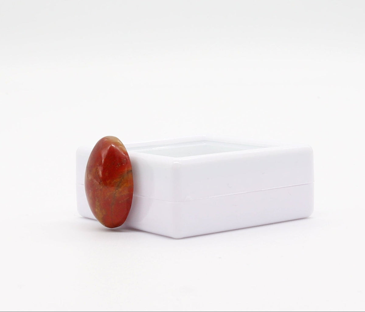 Right angle view of  Sonoran Sunrise Chrysocolla Cuprite gemstone, has been carefully handcut and polished into an oval shape cabochon, revealing its natural vibrant shades of warm reds and yellow, reminiscent of a desert sunrise.  Shown on white background leaning against a white gem gift box.