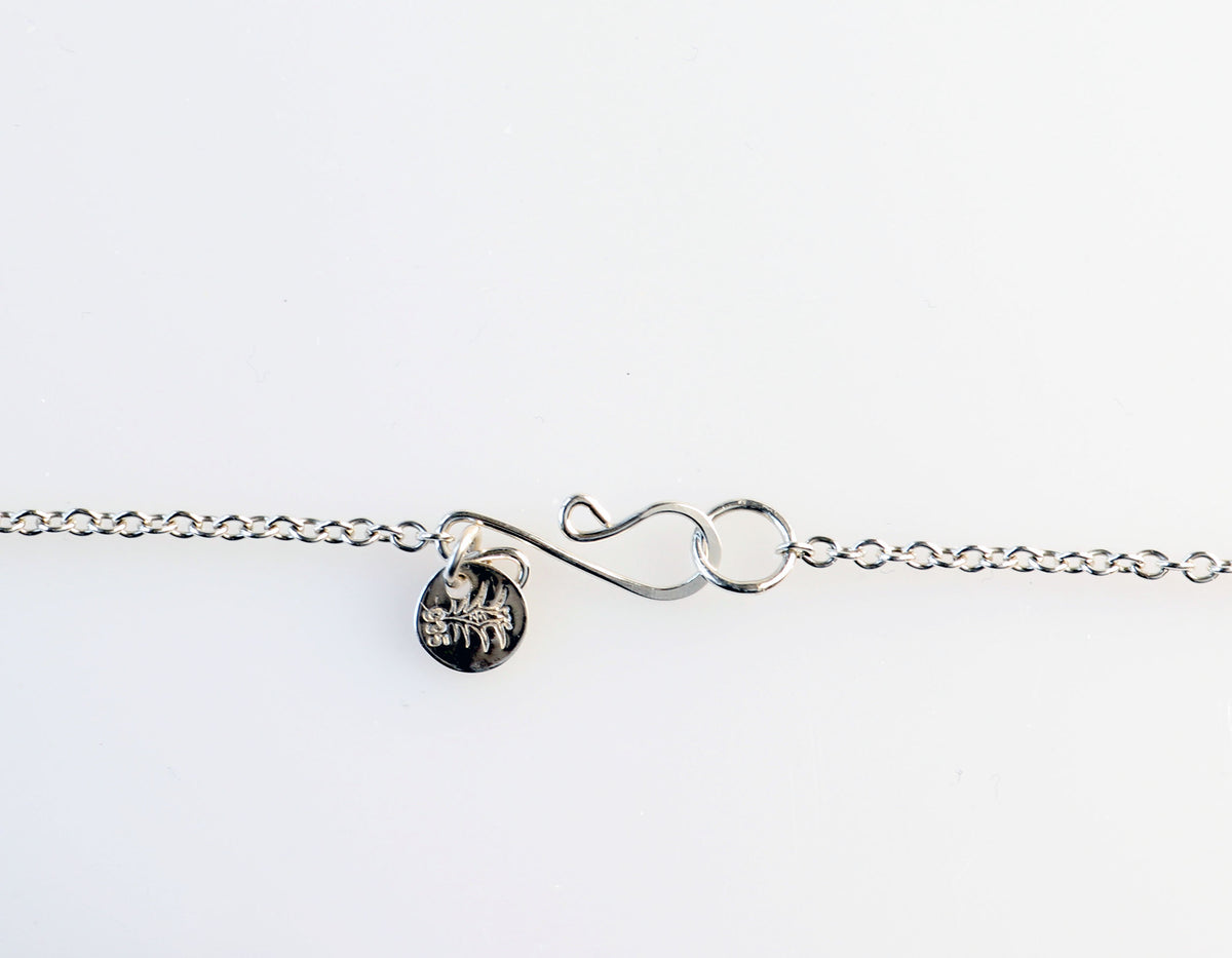 A closeup of the handcrafted custom S-hook clasp in 935 Argentium silver and a small round tag with an engraved makers mark and 935, shown on a plain white background.
